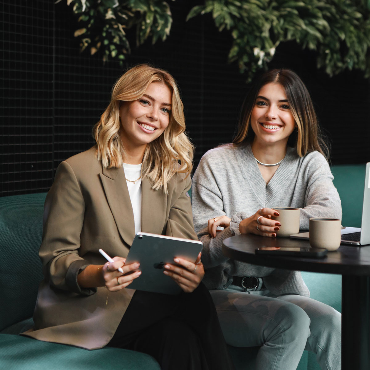 Zwei lächelnde Frauen, eine mit Tablet, die andere mit Kaffeetasse, in einem Café mit Pflanzen.