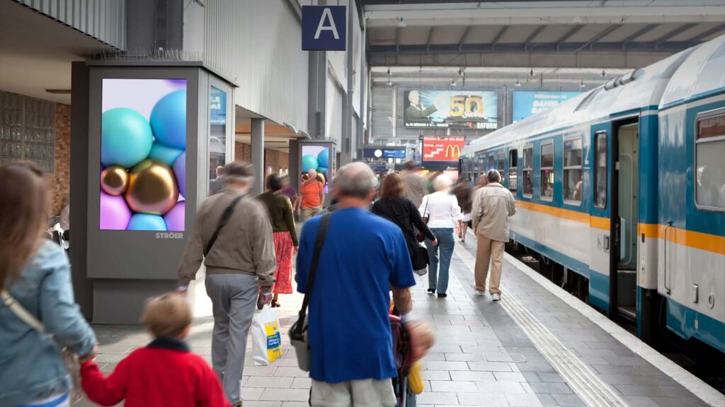 Personen am Bahnhof mit Zug. Farbenfrohes Werbeplakat links. Hektische Stimmung.